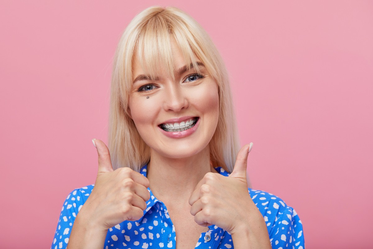 Woman with blonde hair and bangs giving double thumbs up against pink background, wearing blue patterned top.