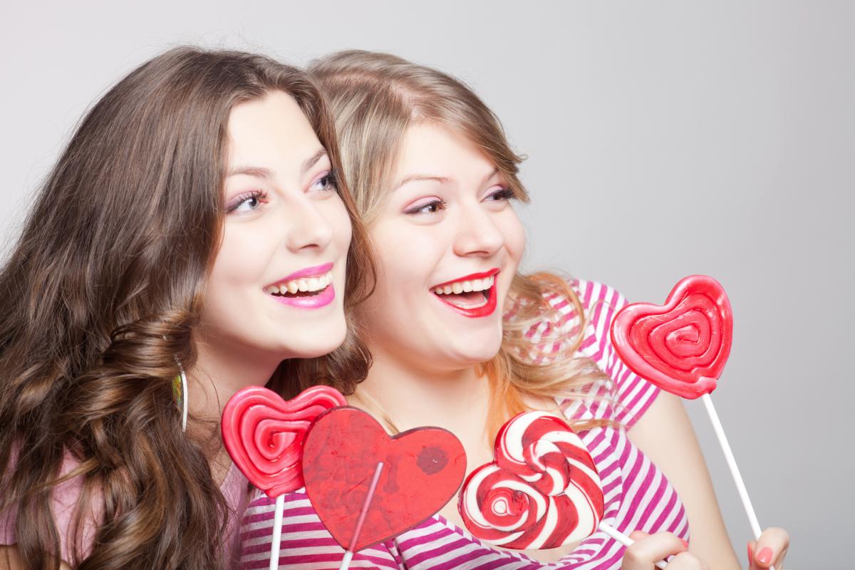 Two smiling women holding pink heart-shaped lollipops against white background.