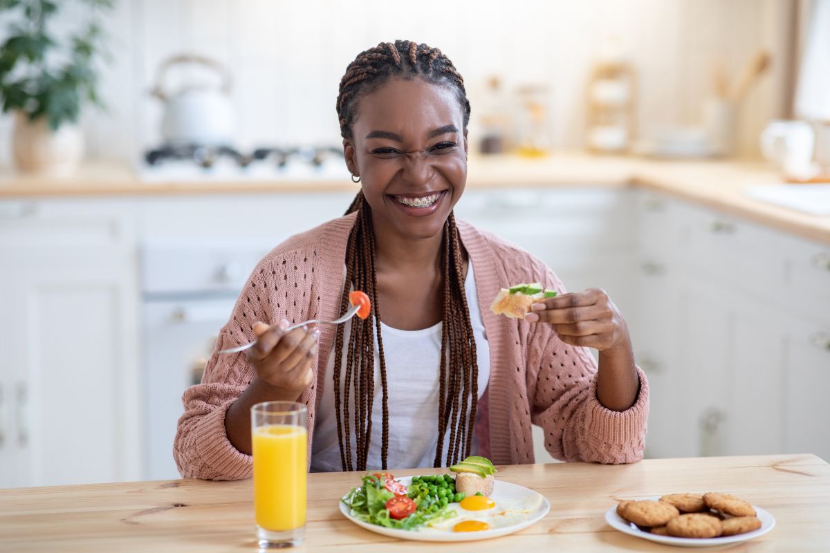 Woman with braids smiling at kitchen table with healthy meal and juice glass.