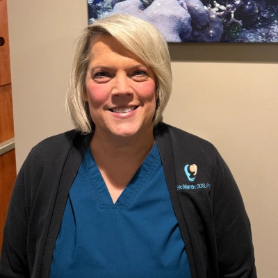 Professional woman in blue scrubs and black cardigan smiling at camera in medical office setting.