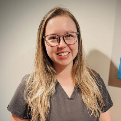 Woman with glasses wearing gray v-neck shirt smiling at camera against white wall