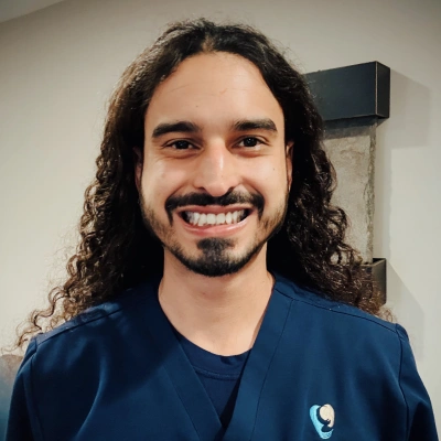 Man with long dark curly hair smiling at camera in casual blue shirt indoors.